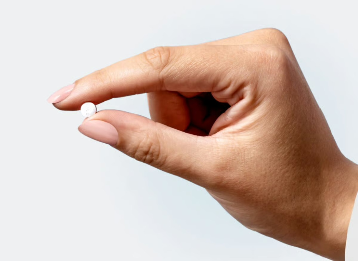 A hand pinches a small white oral GLP-1 tablet between thumb and forefinger against a soft gray background.